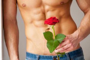 Surprise for her. Close-up of young muscular man with perfect torso holding red rose while leaning at the wall photo