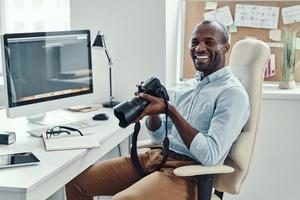 Handsome young African man holding digital camera and smiling while working in the modern office photo
