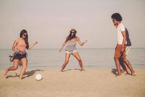 juegos activos con amigos. grupo de jóvenes alegres jugando con una pelota de fútbol en la playa con el mar al fondo foto