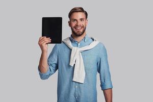 Using technologies.  Good looking young man carrying digital tablet and looking at camera with smile while standing against grey background photo