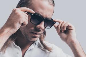 Trendy look. Close-up of handsome young man in white shirt adjusting his sunglasses while standing against grey background photo