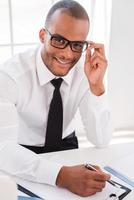 Confident businessman at work. Top view of confident young African man in shirt and tie writing something at clipboard and looking at camera while sitting at his working place photo