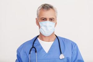 cirujano confiado. retrato de un médico senior de cabello gris con mascarilla quirúrgica mirando a la cámara mientras está de pie aislado en blanco foto