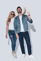 Playful couple. Full length of beautiful young couple in denim wear making peace sign while standing against grey background photo