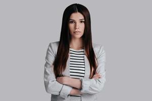 Confident and beautiful. Attractive young woman in smart casual wear keeping arms crossed and looking at camera while standing against grey background photo