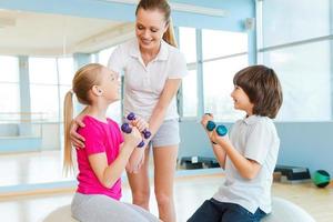 Supporting kids in training. Cheerful instructor helping children with exercising in health club photo