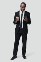 Full length of modern young African man in formalwear using smart phone and smiling while standing against grey background photo