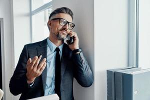 Cheerful businessman talking on mobile phone and gesturing while standing near the window photo