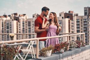 Encantadora pareja joven con ropa informal abrazándose y mirando hacia otro lado con una sonrisa mientras está de pie en el patio de la azotea al aire libre foto