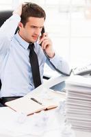 Busy working in office. Worried young man in formalwear talking on the telephone and holding hand in hair while sitting at his working place photo