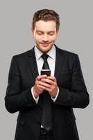 Typing business message. Confident young man in formalwear holding mobile phone and smiling while standing against grey background photo
