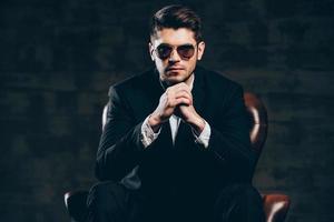 Stylish and successful.Young handsome man in suit and sunglasses keeping hands clasped and looking at camera while sitting in leather chair against dark grey background photo