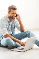 Doing business at home. Smiling young man working on laptop and talking on the mobile phone while sitting on the floor at his apartment photo