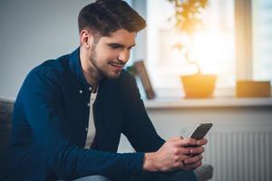 Lovely message from her. Side view of handsome young man using his smartphone with smile while sitting on the couch at home photo