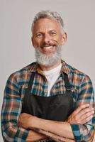 Senior man in apron smiling and looking at camera while standing against gray background photo