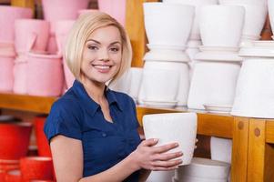 I choose this one pot. Beautiful young blond hair woman holding a flower pot while standing near the shelf with other multi colored pots photo