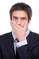 Keeping a secret. Portrait of young man in formalwear covering mouth with hand and looking at camera while standing isolated on white background photo