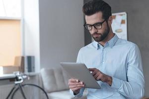 Checking his timetable. Close-up of handsome young man wearing glasses and working with touchpad while standing in office photo