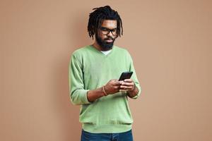 un joven africano pensativo vestido con ropa informal inteligente que usa un teléfono inteligente mientras se enfrenta a un fondo marrón foto