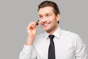 Confident customer service representative. Handsome young man in formalwear and headset looking away and smiling while standing against grey background photo