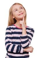 Little daydreamer. Cheerful little girl holding finger on chin and looking away while standing isolated on white photo