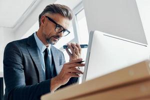 hombre de negocios concentrado que usa un altavoz en el teléfono mientras señala el monitor de la computadora en la oficina foto