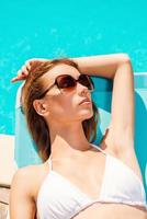el sol es la mejor medicina. vista superior de una hermosa joven en bikini blanco relajándose en una tumbona cerca de la piscina foto