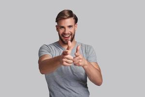 Charming young man pointing at you and smiling while standing against grey background photo