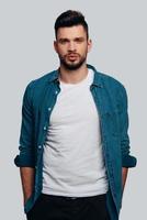 Young and charming. Charming young man looking at camera and keeping hands in pockets while standing against grey background photo