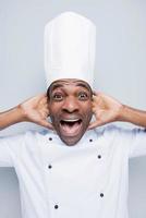 Too much salt Excited young African chef in white uniform touching head with hands and keeping mouth open while standing against grey background photo