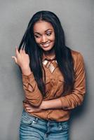 sensual y hermosa. hermosa joven africana manteniendo la mano en el cabello y mirando hacia otro lado con una sonrisa mientras se enfrenta a un fondo gris foto