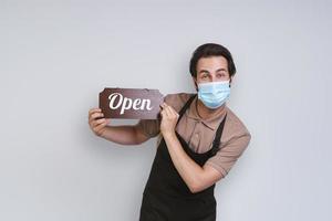 Handsome young man in apron wearing protective face mask and holding open sign while standing against gray background photo