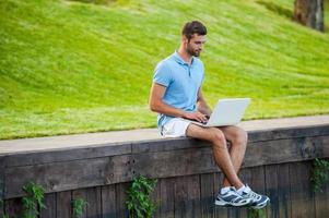 Surfing web wherever he wants. Handsome young man in polo shirt working on laptop while sitting on quayside photo