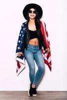 Freedom in in her veins. Full length of beautiful young mixed race woman carrying American flag on shoulders and smiling while standing against white background photo