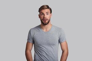 Amazing news. Shocked young man making a face and looking at camera while standing against grey background photo