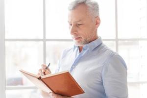 Making some business notes. Serious grey hair senior man in shirt writing something in his note pad and while standing in front of the window photo