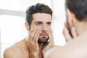 Hangover after night out. Frustrated young shirtless man touching his face and looking at himself while standing against a mirror photo