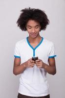 Typing messages to friends. Cheerful African teenager holding mobile phone and looking at it while standing isolated on grey background photo