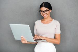 elegante profesional. una joven empresaria sonriente que trabaja en una laptop mientras se enfrenta a un fondo gris foto