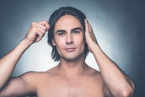 Used to look perfect. Portrait of young shirtless man combing his hair with hairbrush and looking at camera while standing against grey background photo