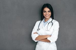 Confident doctor. Attractive young female doctor in white lab coat keeping arms crossed and smiling while standing against grey background photo