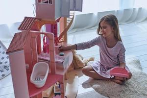 Usando la fantasía para jugar. linda niña jugando con una casa de muñecas mientras se sienta en el suelo en el dormitorio foto