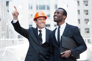 Showing opportunities. Cheerful contractor in hardhat pointing away and smiling while standing together with African businessman photo