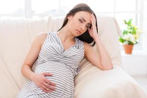 Struggling with morning sickness. Depressed pregnant woman holding hand on head and keeping eyes closed while sitting on a couch photo