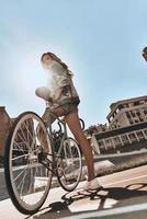 paseo en el sol. longitud completa de una joven atractiva con ropa informal mirando hacia otro lado mientras está de pie cerca de su bicicleta al aire libre con el sol brillando en el fondo foto