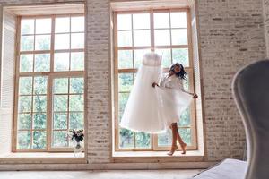 Amazing dress. Full length of beautiful young woman in silk bathrobe touching her wedding dress and smiling while standing near the window photo