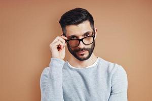 Handsome young man in casual wear looking at camera and adjusting eyewear while standing against brown background photo