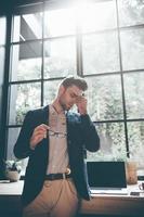 Feeling overworked. Low angle view of frustrated young man carrying eyeglasses and massaging his nose while keeping eyes closed and standing in front of big window in office photo