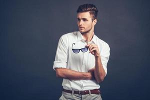 Loving his style. Confident young handsome man carrying his sunglasses and looking away while standing against grey background photo