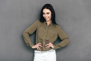 Confident in her look. Happy young woman in smart casual wear standing against grey background and holding hands on hip photo